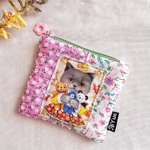 May include: A square, pink floral print coin purse with a green zipper. The center features a gray cat holding teddy bears. The purse has a pink flower-shaped zipper pull and a black tag with white text that reads "AiYue".
