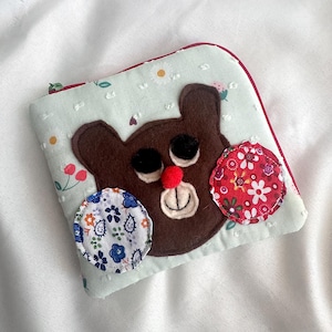 May include: A handmade, light blue zippered pouch with a brown bear face applique. The bear has black eyes, a red nose, and a stitched smile. Decorative floral fabric circles and a red floral trim add detail.