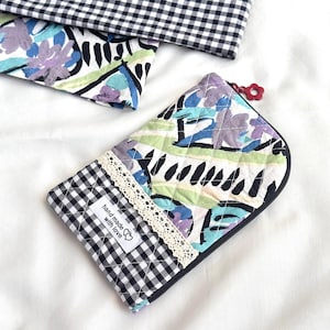 May include: A quilted pouch with a black and white gingham pattern and a floral design in shades of blue, purple, and green. The pouch has a black zipper and a small red flower charm. The label reads "hand made with love".