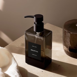 Puede incluir: Una botella de vidrio transparente con un dispensador de bomba negro lleno de loción para manos. La botella tiene una etiqueta negra con el texto "CLEO + SAINT SOLSTICE HAND LOTION".