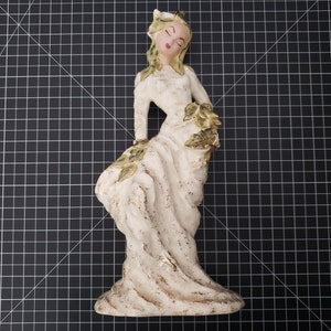 May include: A white ceramic figurine of a woman with green leaves in her hair and dress. The figurine is standing on a black and white grid background.