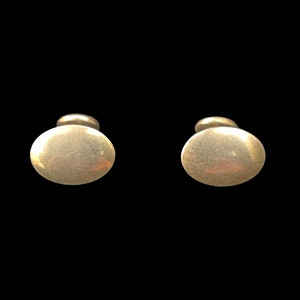 May include: Two gold-colored cufflinks with a simple, oval design. The cufflinks have a polished finish and a classic, understated look. The background is solid black, highlighting the jewelry.