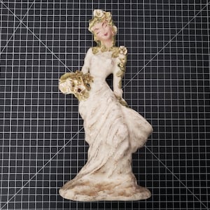 May include: A white ceramic figurine of a woman wearing a white dress and a floral crown. She is holding a bouquet of flowers in her left hand and her right hand is on her hip. The figurine is standing on a base with a textured surface.