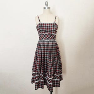 May include: A sleeveless midi dress featuring a red, black, and white plaid pattern. The garment has thin shoulder straps, a fitted bodice, and a flared skirt. White lace trim accents the neckline, waist, and two horizontal rows on the skirt.