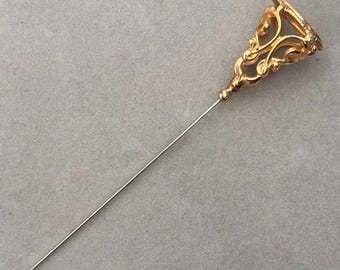 May include: A gold-colored hat pin with an ornate, openwork design at the top. The pin has a long, slender, silver-colored shaft and a decorative, bell-shaped head. The head features intricate scrollwork and a small, round detail.