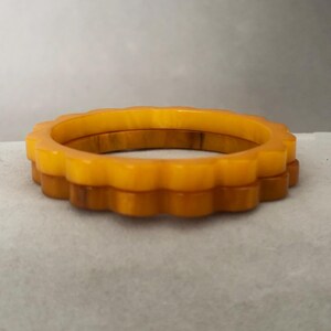 May include: Two vintage, amber-colored bangles with a scalloped edge. The bracelets are made of a translucent material, with a darker amber hue on the bottom bangle. The bangles are stacked on top of each other.