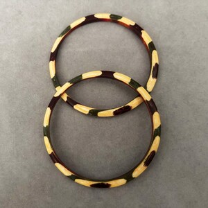 May include: Two round bangles with a camouflage pattern in shades of brown, green, and cream. The bangles are interlocked, creating an overlapping design. The jewelry is set against a gray background.