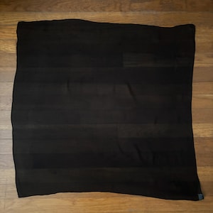 May include: A solid black scarf, rectangular in shape, is displayed against a wooden background. The scarf appears to be made of a lightweight fabric, with subtle texture visible. The dark color suggests versatility for various outfits.