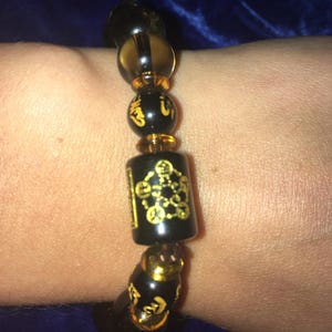 May include: A black beaded bracelet with gold accents. The bracelet features a cylindrical charm with a gold design. The beads are interspersed with amber-coloured beads.