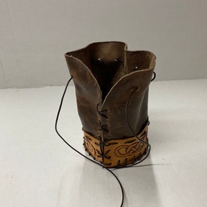 May include: A brown leather pouch with a drawstring closure. The base of the pouch has a lighter brown band with a Celtic knot design. A black cord is attached for carrying.