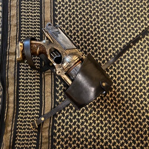 Pode incluir: Uma pistola de estilo steampunk com uma bainha de couro marrom e uma alça preta. A pistola é feita de metal e tem um acabamento dourado. A bainha é presa à alça com uma fivela de metal.