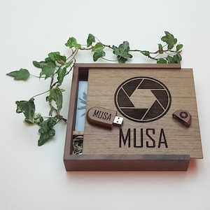 May include: A wooden box with a dark brown finish, engraved with the word "MUSA" and a camera shutter design. The box contains a small wooden USB drive with the word "MUSA" engraved on it.