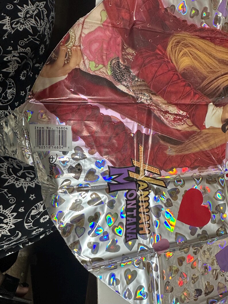 Puede incluir: Un globo de papel de aluminio plateado metalizado con un gr&aacute;fico de Hannah Montana con chaqueta roja y pelo rubio. La superficie est&aacute; decorada con corazones hologr&aacute;ficos e incluye el texto Hannah Montana, un logo de Disney y un c&oacute;digo de barras n.&ordm; 16804.