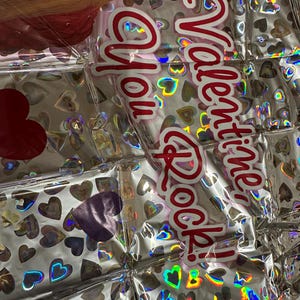 Puede incluir: Un globo de papel de aluminio plateado con un patr&oacute;n repetitivo de peque&ntilde;os corazones hologr&aacute;ficos y el texto rojo Valentine, You Rock! La superficie del globo es met&aacute;lica y reflectante, mostrando una imagen parcial de una persona con un micr&oacute;fono.