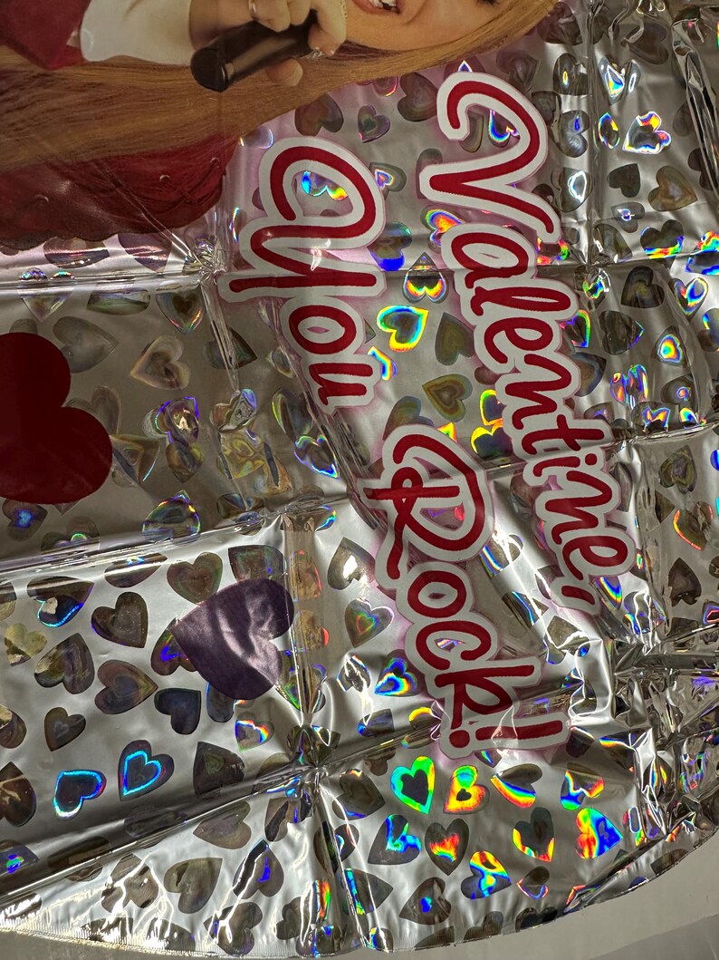 Puede incluir: Un globo de papel de aluminio plateado con un patr&oacute;n repetitivo de peque&ntilde;os corazones hologr&aacute;ficos y el texto rojo Valentine, You Rock! La superficie del globo es met&aacute;lica y reflectante, mostrando una imagen parcial de una persona con un micr&oacute;fono.