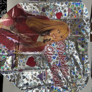 Puede incluir: Un globo de papel de aluminio plateado con un patr&oacute;n de corazones hologr&aacute;ficos y una imagen impresa de Hannah Montana sosteniendo un micr&oacute;fono. El globo incluye el texto rojo "Valentine, You Rock!" y el logotipo de Hannah Montana en letras doradas y negras.