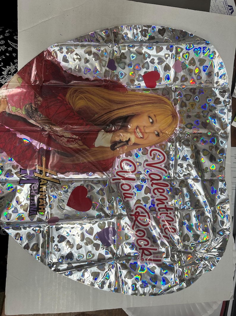 Puede incluir: Un globo de papel de aluminio plateado con un patr&oacute;n de corazones hologr&aacute;ficos y una imagen impresa de Hannah Montana sosteniendo un micr&oacute;fono. El globo incluye el texto rojo "Valentine, You Rock!" y el logotipo de Hannah Montana en letras doradas y negras.