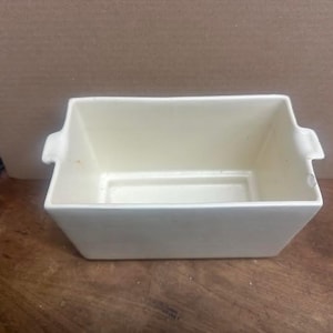 Puede incluir: Una fuente rectangular de cerámica blanquecina con dos pequeñas asas en los laterales. La fuente tiene un diseño ligeramente cónico y se apoya sobre una superficie de madera. El interior es de un tono más claro de blanquecino.