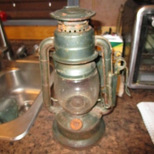 May include: A vintage green kerosene lantern with a glass globe and a metal handle. The lantern has a brass-colored top and a rusty base.