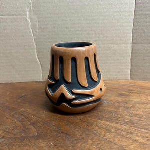 May include: A small, handcrafted ceramic vase with a unique design. The vase features a brown exterior with a black interior and intricate cut-out patterns. The vase is approximately 3 inches tall and has a cylindrical shape.