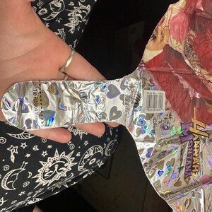 Puede incluir: Un globo de aluminio con un gr&aacute;fico de Hannah Montana y un patr&oacute;n de corazones hologr&aacute;ficos. El globo tiene una leng&uuml;eta plateada con el texto 'Tie Ribbon Here' y una etiqueta de c&oacute;digo de barras 'Item No. 16804' cerca del logo de Disney.