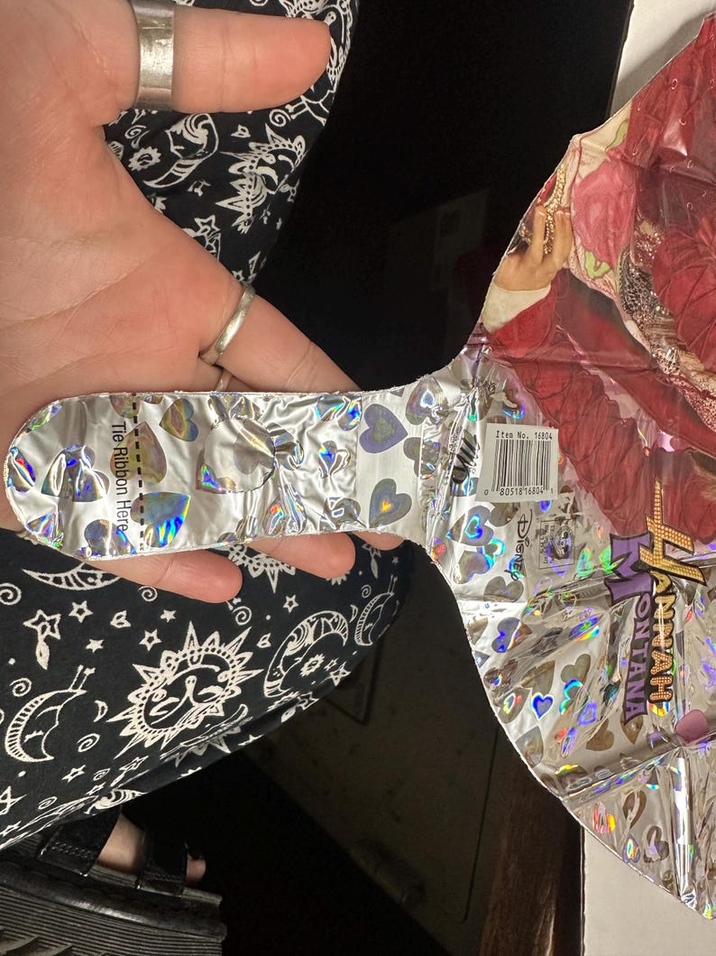 Puede incluir: Un globo de aluminio con un gr&aacute;fico de Hannah Montana y un patr&oacute;n de corazones hologr&aacute;ficos. El globo tiene una leng&uuml;eta plateada con el texto 'Tie Ribbon Here' y una etiqueta de c&oacute;digo de barras 'Item No. 16804' cerca del logo de Disney.