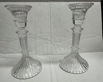 Könnte beinhalten: Zwei Kerzenhalter aus klarem Glas. Jeder hat einen ausgestellten Sockel mit geripptem Design, einen schlanken, geriffelten Stiel und eine breite, geriffelte Oberseite. Die Kerzenhalter sind identisch und scheinen aus Kristall oder einem ähnlichen Material zu bestehen.
