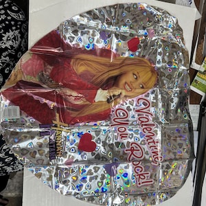 Puede incluir: Un globo de papel de aluminio plateado con un patr&oacute;n de corazones hologr&aacute;ficos y una imagen impresa de Hannah Montana. El globo muestra el texto "Valentine You Rock!" en letras rojas e incluye el logotipo de Hannah Montana en morado y dorado.
