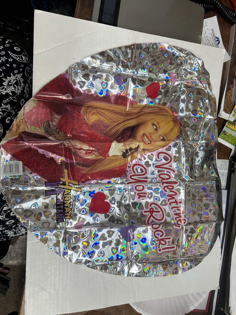 Puede incluir: Un globo de papel de aluminio plateado con un patr&oacute;n de corazones hologr&aacute;ficos y una imagen impresa de Hannah Montana. El globo muestra el texto "Valentine You Rock!" en letras rojas e incluye el logotipo de Hannah Montana en morado y dorado.