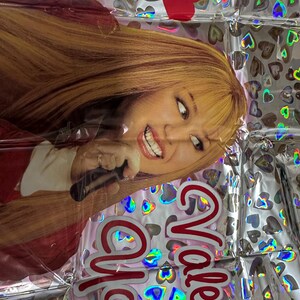 Puede incluir: Un globo de aluminio rectangular con una impresi&oacute;n fotogr&aacute;fica de una persona con cabello rubio largo sosteniendo un micr&oacute;fono. El fondo es plateado con un patr&oacute;n repetitivo de corazones hologr&aacute;ficos. El texto "Valentine You" est&aacute; en rojo.