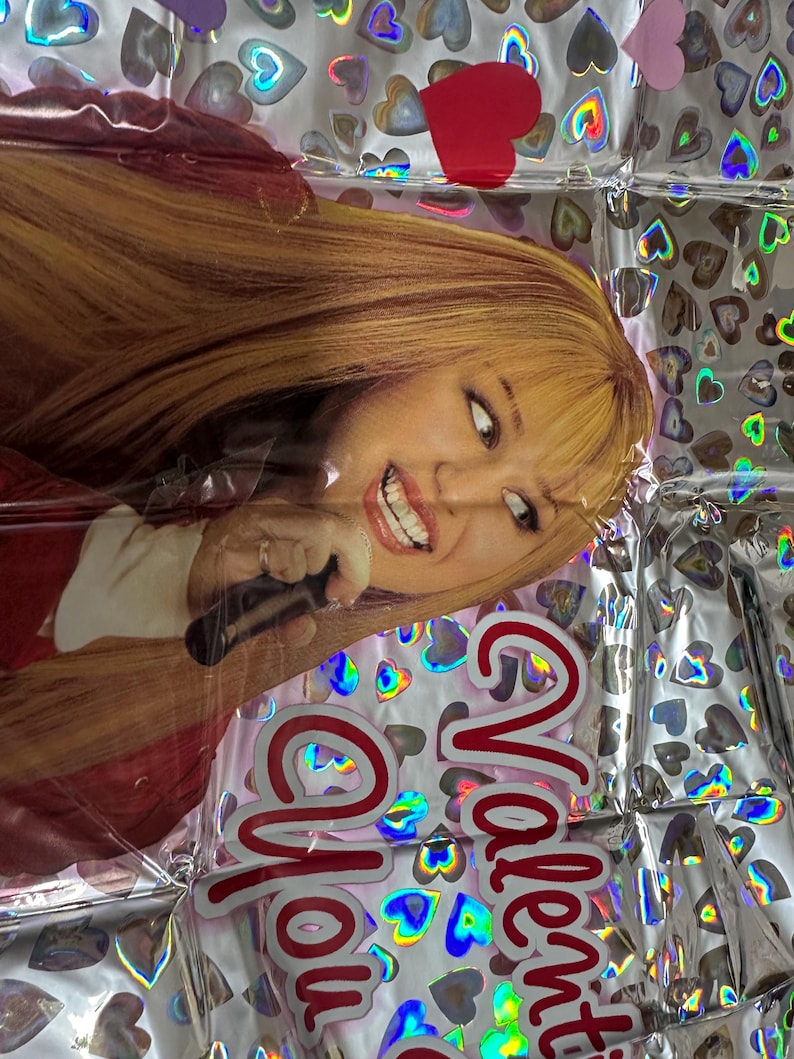 Puede incluir: Un globo de aluminio rectangular con una impresi&oacute;n fotogr&aacute;fica de una persona con cabello rubio largo sosteniendo un micr&oacute;fono. El fondo es plateado con un patr&oacute;n repetitivo de corazones hologr&aacute;ficos. El texto "Valentine You" est&aacute; en rojo.