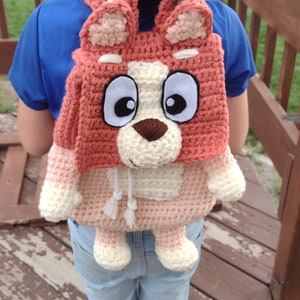 May include: A crocheted backpack shaped like a cartoon dog with pink, white, and brown fur. The backpack has large eyes, a brown nose, and a white drawstring.