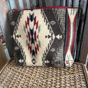 May include: A rectangular, zippered pouch with a Southwestern-inspired pattern. The design features a cream-colored central panel with a geometric pattern in shades of brown, red, teal, and navy. The sides of the pouch are a darker brown with a similar pattern.