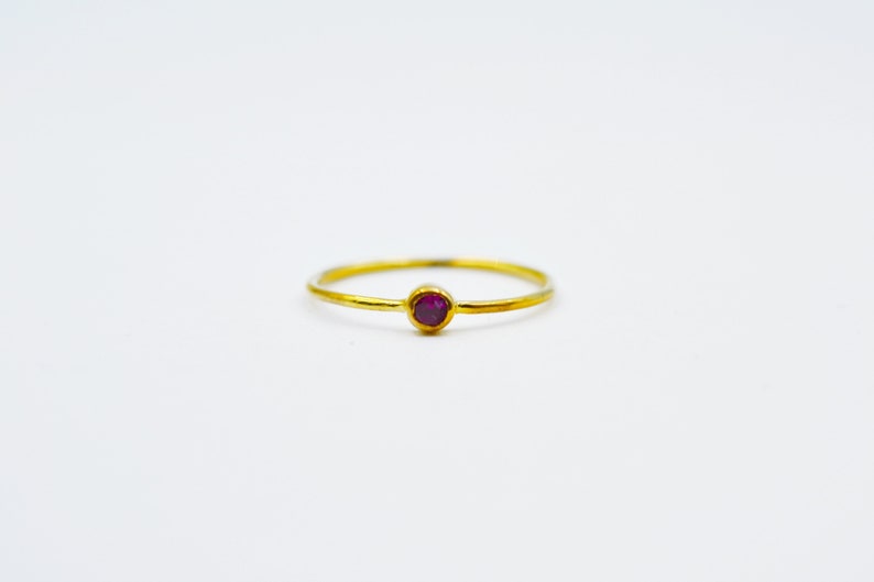 以下が含まれることがあります： 小さな丸い濃い赤色の宝石が付いたシンプルなゴールドバンドリングです。