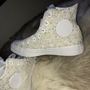 May include: White high-top sneakers covered in white pearl embellishments. The sneakers have a white star logo on the side.