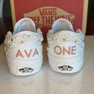 Puede incluir: Un par de Vans blancas de deslizamiento con adornos de perlas. Un zapato dice "AVA" y el otro dice "ONE" en letras doradas.