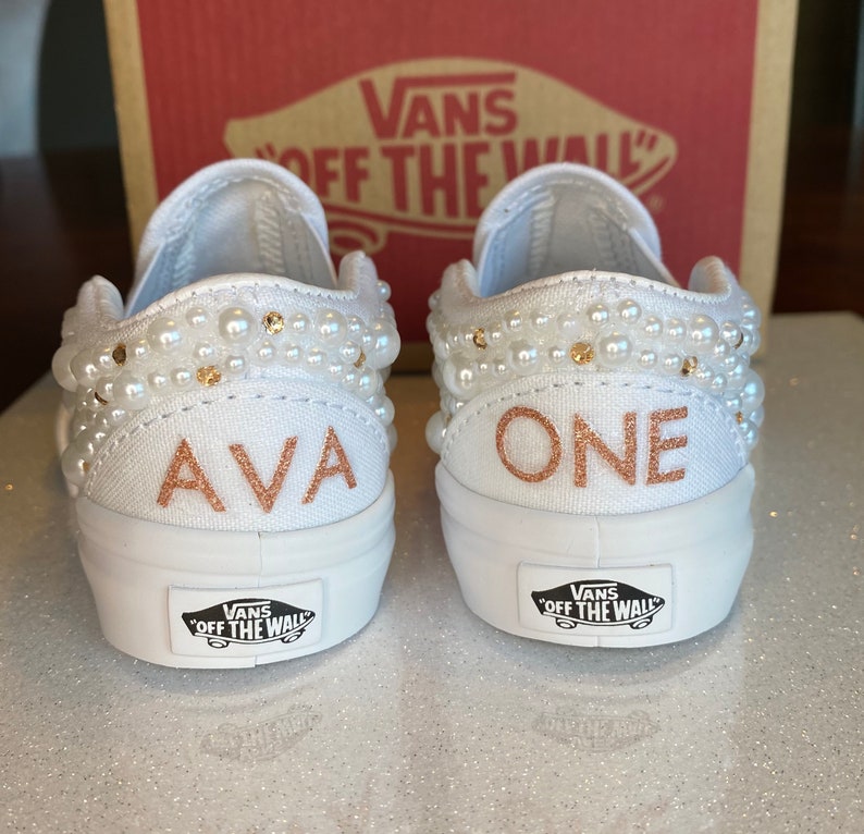 Puede incluir: Un par de Vans blancas de deslizamiento con adornos de perlas. Un zapato dice "AVA" y el otro dice "ONE" en letras doradas.