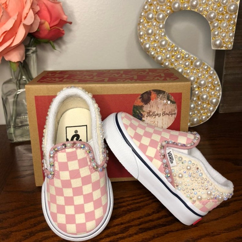 Puede incluir: Zapatillas Vans de slip-on a cuadros rosa y blanco con adornos de perlas y diamantes de imitaci&oacute;n. Los zapatos est&aacute;n decorados con un borde de perlas alrededor del borde y tienen una suela blanca.