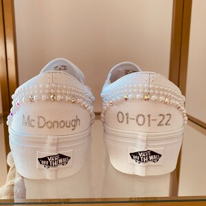 Puede incluir: Un par de zapatillas blancas de lona sin cordones con adornos de perlas. El zapato izquierdo tiene el texto "Mc Donough" y el zapato derecho tiene la fecha "01-01-22" escrita en ellos. Los zapatos tienen el logotipo de Vans en la parte posterior.