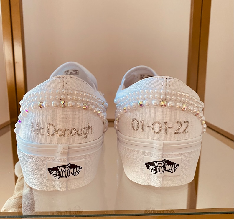 Puede incluir: Un par de zapatillas blancas de lona sin cordones con adornos de perlas. El zapato izquierdo tiene el texto "Mc Donough" y el zapato derecho tiene la fecha "01-01-22" escrita en ellos. Los zapatos tienen el logotipo de Vans en la parte posterior.