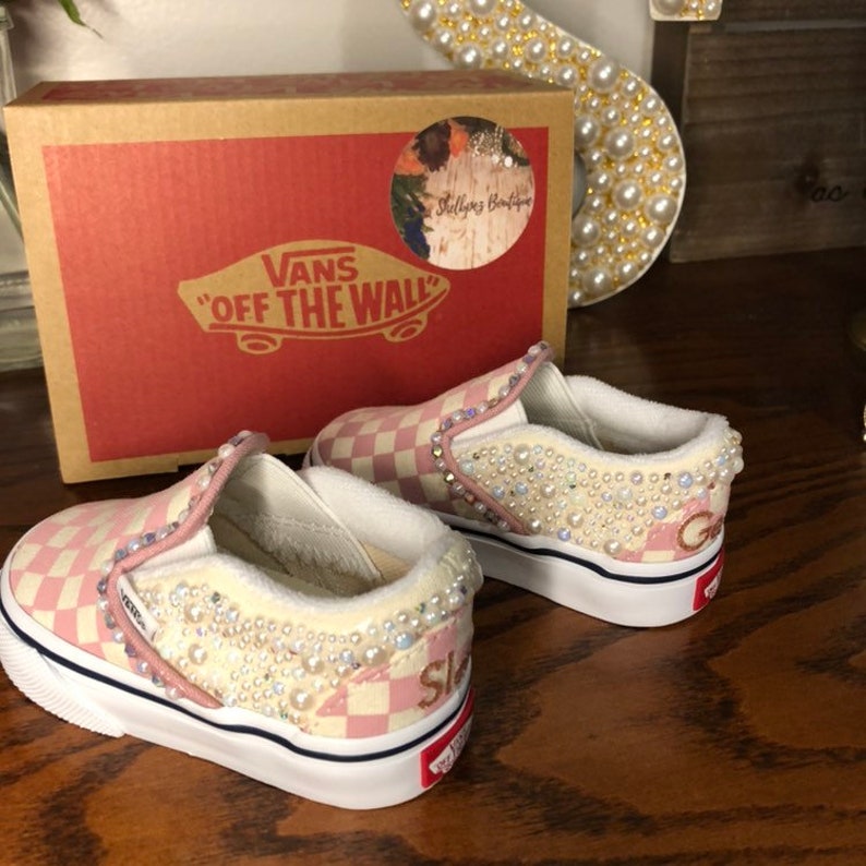 Puede incluir: Un par de zapatos Vans slip-on a cuadros rosa y blanco con adornos de perlas. Los zapatos est&aacute;n decorados con pedrer&iacute;a y perlas, y el nombre de la marca "Vans" y "Off The Wall" son visibles en el lateral de los zapatos.