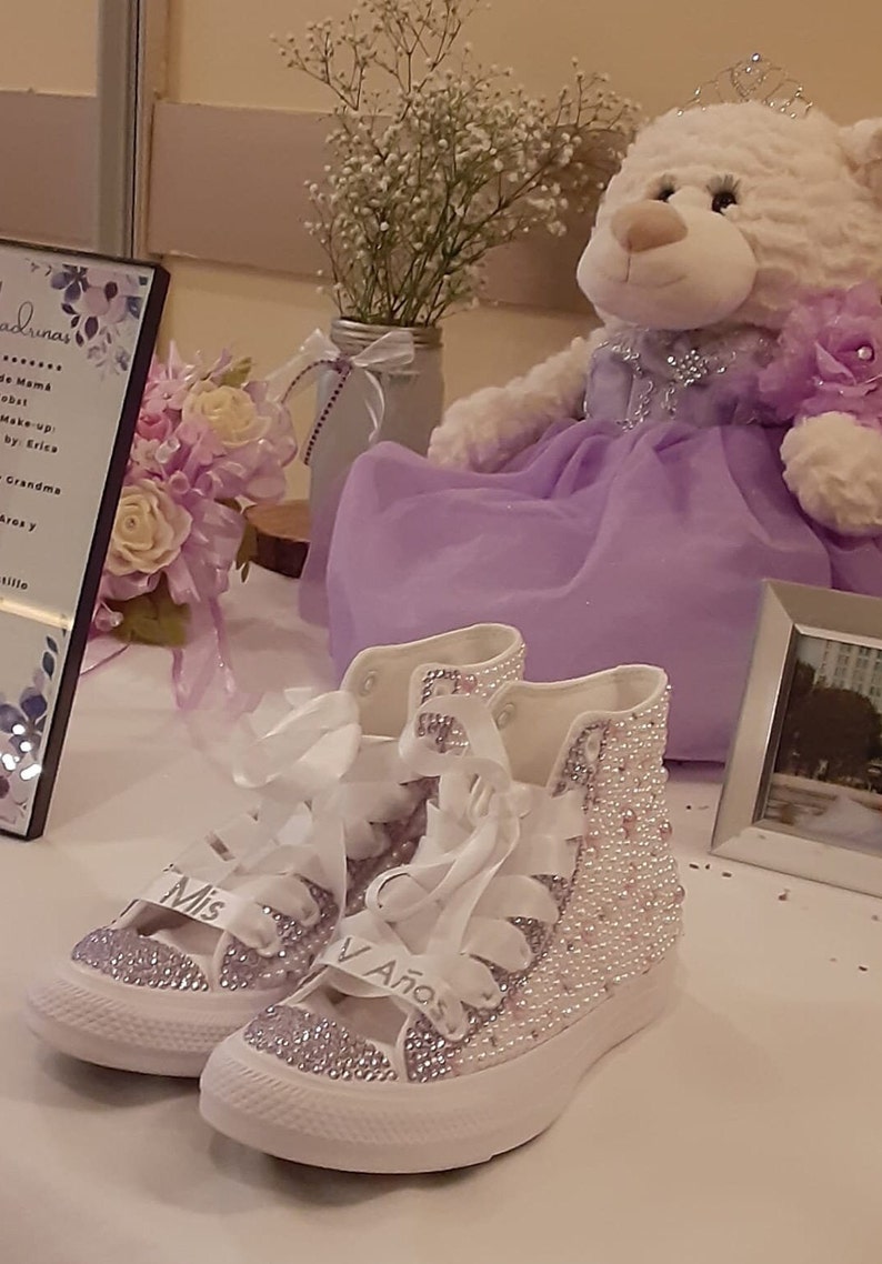 May include: A pair of white high-top sneakers with a purple and white design. The sneakers are covered in rhinestones and have the words "Mis 15 A&ntilde;os" written on them.