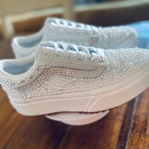 Könnte beinhalten: Weiße Canvas-Sneaker mit einem weißen Perlen- und Strass-Design. Die Schuhe haben eine weiße Gummisohle.
