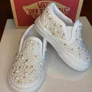 Puede incluir: Un par de Vans blancas de deslizamiento cubiertas de perlas blancas. Los zapatos tienen un dise&ntilde;o cl&aacute;sico de Vans con una suela de goma blanca y una parte superior de lona blanca. Las perlas est&aacute;n distribuidas uniformemente en toda la superficie de los zapatos, creando un aspecto brillante y elegante.