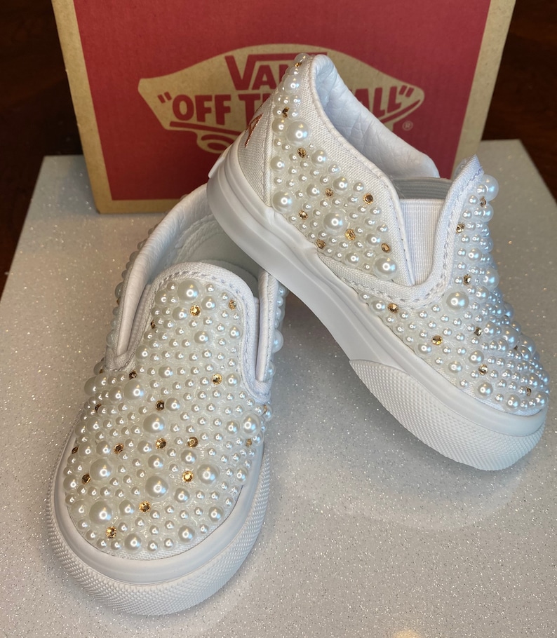 Puede incluir: Un par de Vans blancas de deslizamiento cubiertas de perlas blancas. Los zapatos tienen un dise&ntilde;o cl&aacute;sico de Vans con una suela de goma blanca y una parte superior de lona blanca. Las perlas est&aacute;n distribuidas uniformemente en toda la superficie de los zapatos, creando un aspecto brillante y elegante.