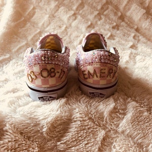 Puede incluir: Un par de zapatillas Vans a cuadros rosa y blanco con adornos de perlas. El zapato izquierdo tiene la fecha "08-08-17" escrita en letras doradas, y el zapato derecho tiene el nombre "EMERY" escrito en letras doradas.