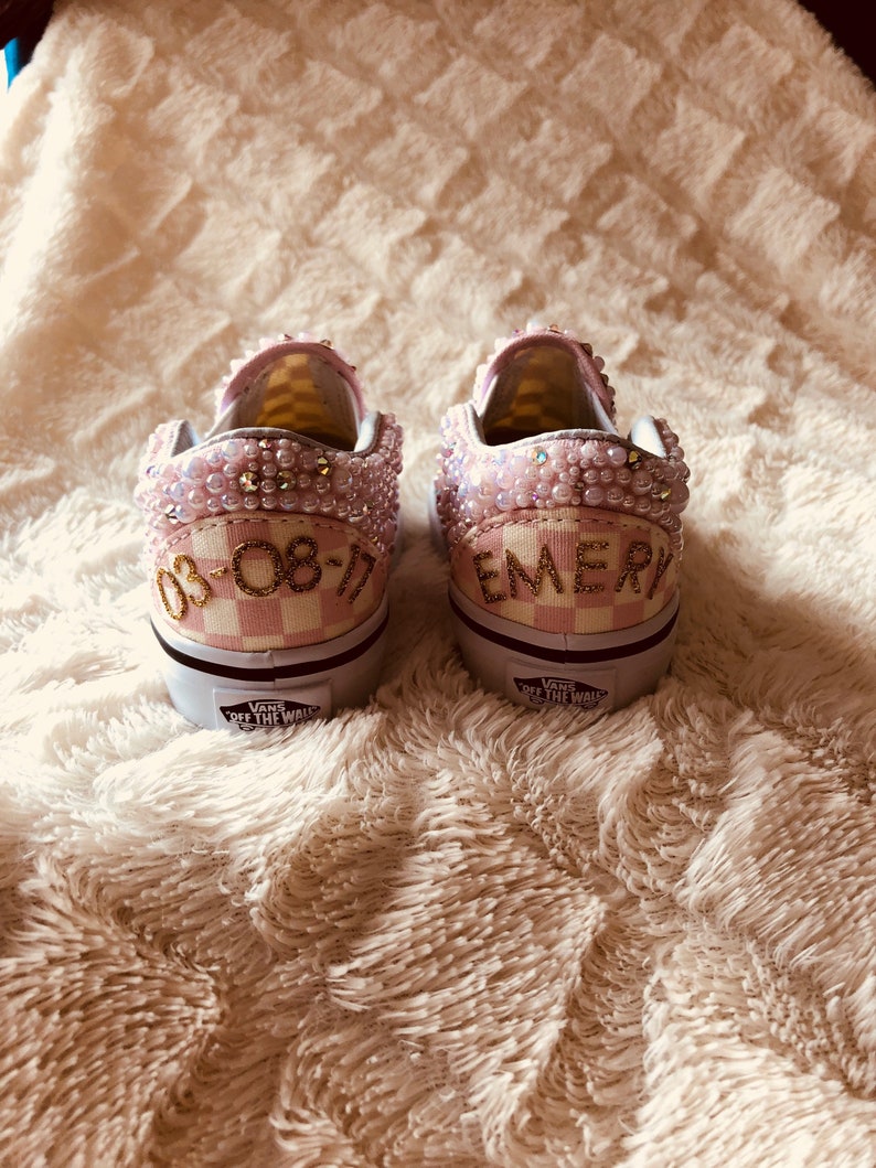 Puede incluir: Un par de zapatillas Vans a cuadros rosa y blanco con adornos de perlas. El zapato izquierdo tiene la fecha "08-08-17" escrita en letras doradas, y el zapato derecho tiene el nombre "EMERY" escrito en letras doradas.