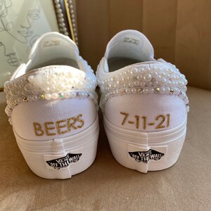 Puede incluir: Zapatillas Vans blancas de slip-on con ribete de perlas y letras doradas. Un zapato dice "BEERS" y el otro dice "7-11-21".