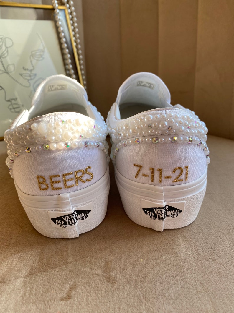 Puede incluir: Zapatillas Vans blancas de slip-on con ribete de perlas y letras doradas. Un zapato dice "BEERS" y el otro dice "7-11-21".