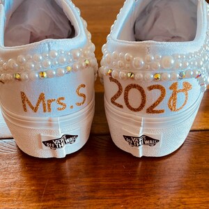 Puede incluir: Un par de zapatos de lona blancos con adornos de perlas. Un zapato dice "Mrs. S" y el otro dice "2023".